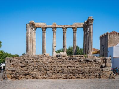 A wide-angle photograph of the Roman Temple of Évora, showing its six Corinthian columns standing on a high stone podium within a paved public square under a clear blue sky.