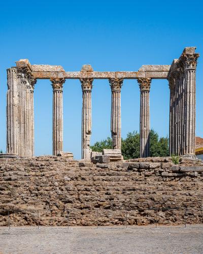 A wide-angle photograph of the Roman Temple of Évora, showing its six Corinthian columns standing on a high stone podium within a paved public square under a clear blue sky.