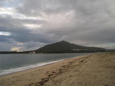 A wide sandy beach with driftwood in the foreground, a body of water to the left, and a large, conical, forested hill rising in the background under a cloudy sky.