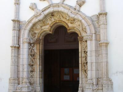 A close-up photograph of an ornate stone church portal featuring Manueline architectural elements, including twisted columns, a cross of the Order of Christ, and a heart-shaped tympanum, set within a white-washed wall.