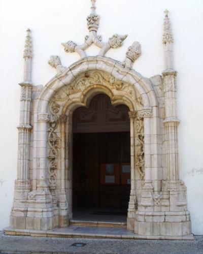 A close-up photograph of an ornate stone church portal featuring Manueline architectural elements, including twisted columns, a cross of the Order of Christ, and a heart-shaped tympanum, set within a white-washed wall.
