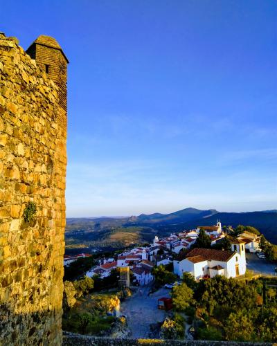 A view from a stone fortification looking down onto a white-washed village with terracotta roofs nestled on a hillside, with rolling hills in the background under a blue sky.