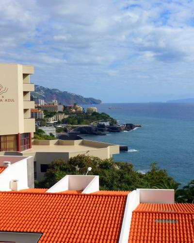 A coastal view of Funchal, Portugal, featuring orange-tiled rooftops in the foreground, the Hotel Baia Azul building on the left, and the Atlantic Ocean stretching to the horizon under a partly cloudy sky.