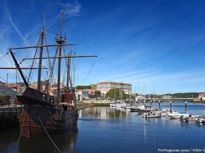 A low-angle shot of a large, dark wooden replica of a 16th-century carrack docked in a harbor, with a modern grandstand and town buildings visible in the background.