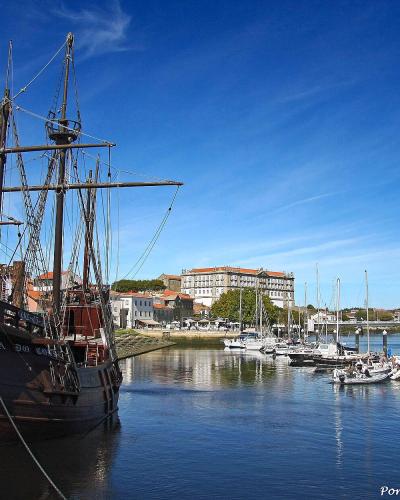 A low-angle shot of a large, dark wooden replica of a 16th-century carrack docked in a harbor, with a modern grandstand and town buildings visible in the background.