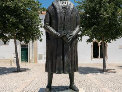 A bronze statue of a man in historical robes standing on a stone pedestal in a paved courtyard, with a white building featuring arched windows and a green door in the background.