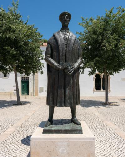 A bronze statue of a man in historical robes standing on a stone pedestal in a paved courtyard, with a white building featuring arched windows and a green door in the background.