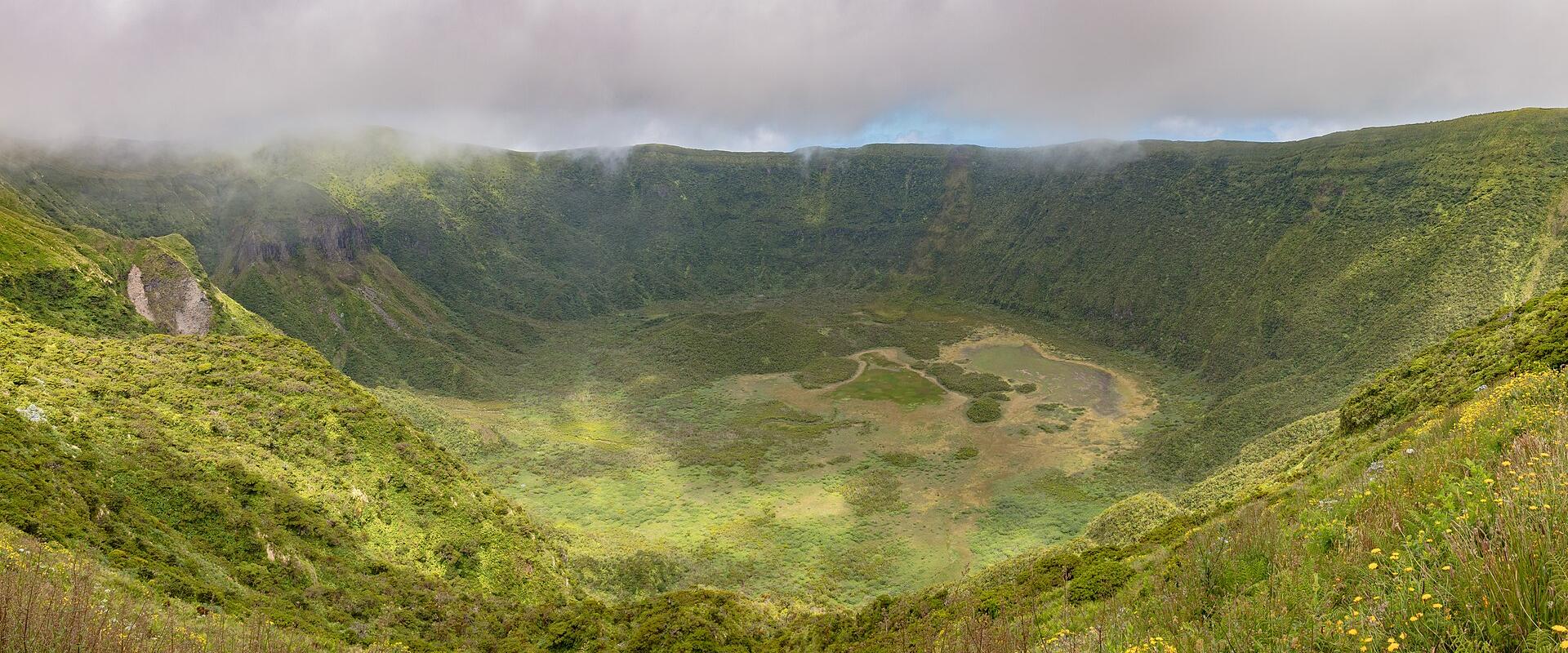 A panoramic view of a large, green volcanic crater (caldera) with a marshy floor and misty clouds hovering above the rim.