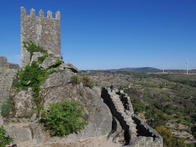 A stone medieval tower and fortified wall built upon a rocky outcrop, overlooking a rural landscape with wind turbines in the distance.