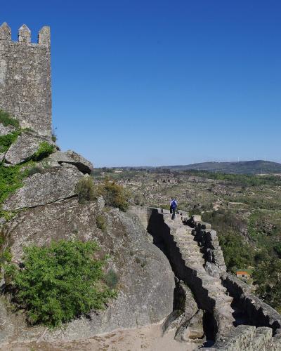 A stone medieval tower and fortified wall built upon a rocky outcrop, overlooking a rural landscape with wind turbines in the distance.