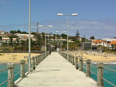 A concrete pier or jetty extending into turquoise water, lined with tall modern streetlights and concrete railings, leading towards a sandy beach and a town with white buildings and orange roofs in the background.