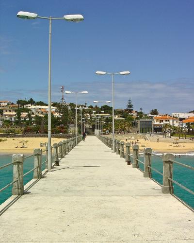 A concrete pier or jetty extending into turquoise water, lined with tall modern streetlights and concrete railings, leading towards a sandy beach and a town with white buildings and orange roofs in the background.