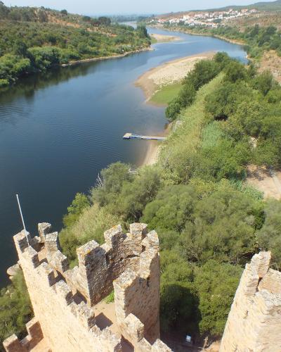 A high-angle view from the battlements of a medieval stone castle overlooking a wide river (the Tagus) with a small islet, dense green vegetation, and a distant town on the far bank; a flagpole is visible on the castle wall.