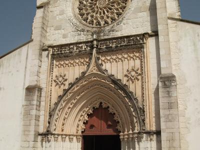 A low-angle photograph of the stone facade of a church featuring a large rose window and an ornate, pointed Gothic entrance portal.