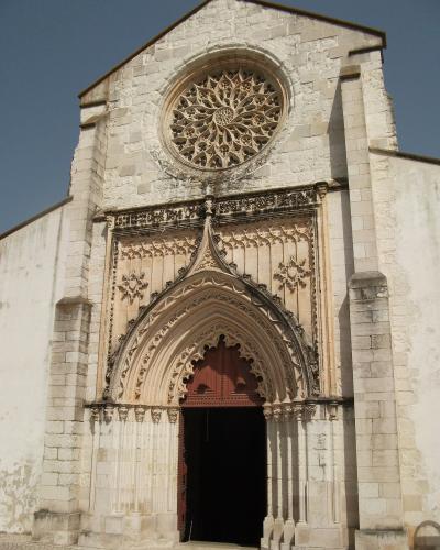 A low-angle photograph of the stone facade of a church featuring a large rose window and an ornate, pointed Gothic entrance portal.