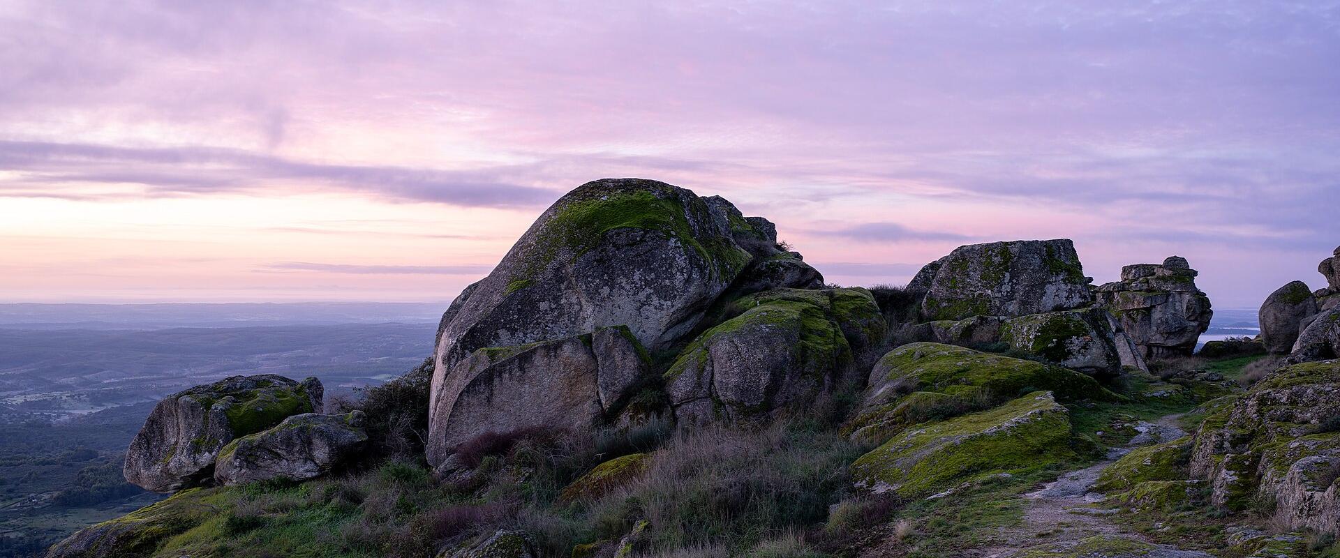 A landscape photograph showing large, moss-covered granite boulders on a hilltop at sunrise or sunset, with a view of a valley below.