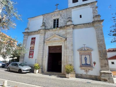 A low-angle photograph of a white church facade with a stone-trimmed entrance, a bell tower, and a red banner hanging on the left wall.