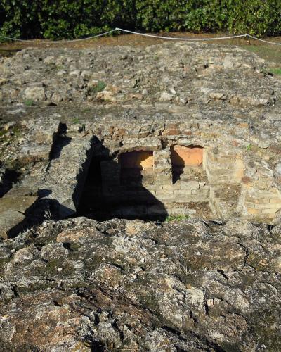 The image shows a close-up view of ancient stone ruins featuring two rectangular niches set into a low wall, surrounded by rough, weathered rock and grass, with a rope barrier visible in the background.