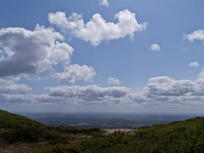 A landscape photograph taken from a high vantage point looking out over a vast, flat plain under a blue sky with scattered white clouds, with green scrubby vegetation in the immediate foreground.