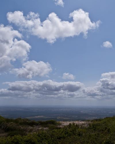 A landscape photograph taken from a high vantage point looking out over a vast, flat plain under a blue sky with scattered white clouds, with green scrubby vegetation in the immediate foreground.