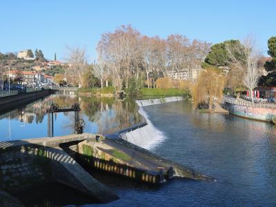 A river scene showing a low weir or small dam with water flowing over it, flanked by a paved walkway with railings on the left and a park area with trees and a riverside café on the right, with a hillside town in the background.