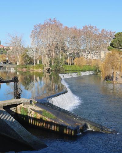A river scene showing a low weir or small dam with water flowing over it, flanked by a paved walkway with railings on the left and a park area with trees and a riverside café on the right, with a hillside town in the background.