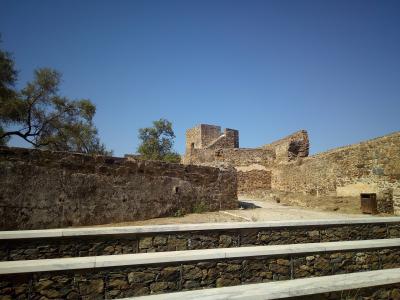 The image shows the stone ruins of a medieval fortress with a square tower and high walls, viewed from behind a modern gabion retaining wall with marble coping under a clear blue sky.