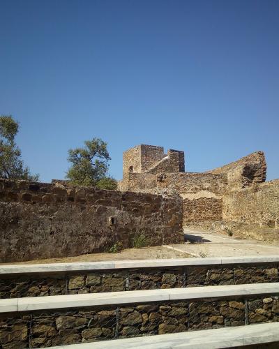 The image shows the stone ruins of a medieval fortress with a square tower and high walls, viewed from behind a modern gabion retaining wall with marble coping under a clear blue sky.