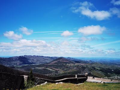 A view from a grassy hilltop featuring a low stone wall in the foreground, overlooking a vast landscape of rolling hills and mountains under a blue sky with clouds.