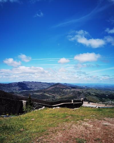 A view from a grassy hilltop featuring a low stone wall in the foreground, overlooking a vast landscape of rolling hills and mountains under a blue sky with clouds.