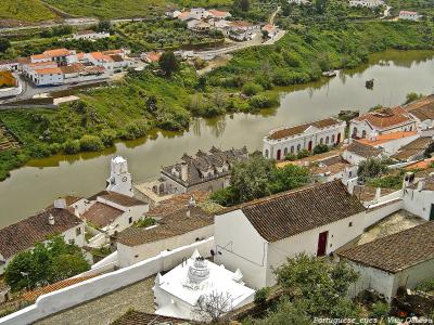 An elevated view of a riverside town featuring white-washed buildings with terracotta roofs, a prominent church with a bell tower, and a river with boats in the background.