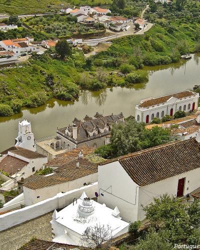 An elevated view of a riverside town featuring white-washed buildings with terracotta roofs, a prominent church with a bell tower, and a river with boats in the background.