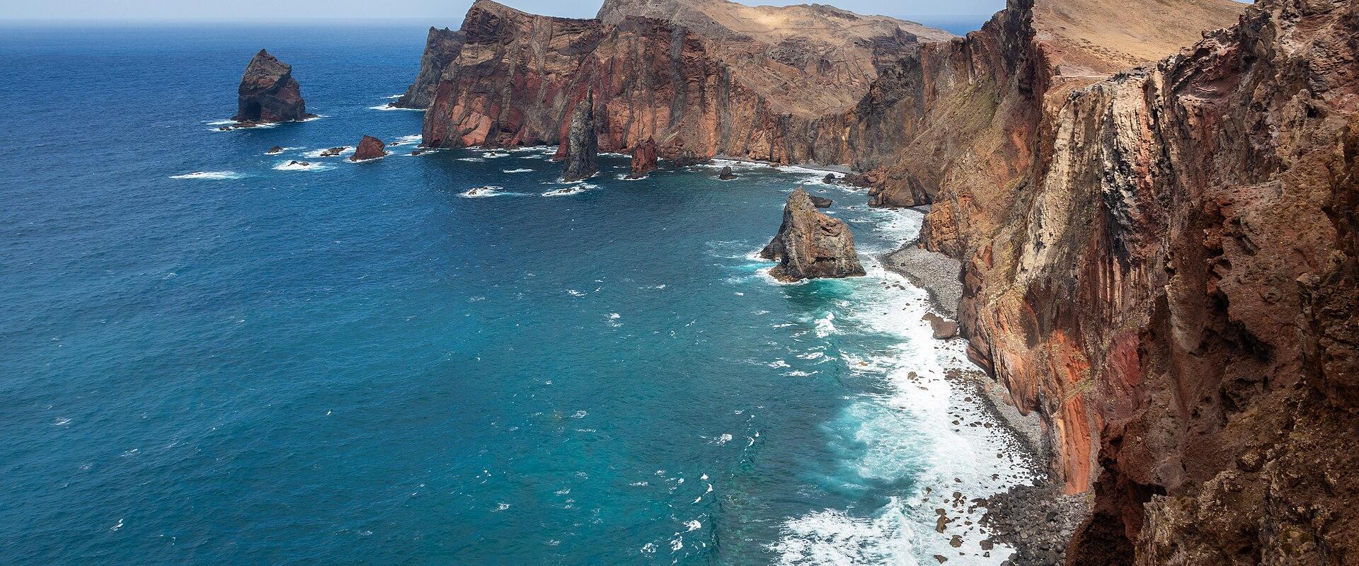 A high-angle view of a rugged, volcanic coastline with steep, reddish-brown cliffs dropping into deep blue ocean water, featuring prominent sea stacks and rocky outcrops along the shore.