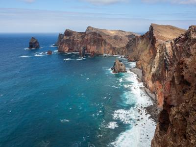 A high-angle view of a rugged, volcanic coastline with steep, reddish-brown cliffs dropping into deep blue ocean water, featuring prominent sea stacks and rocky outcrops along the shore.