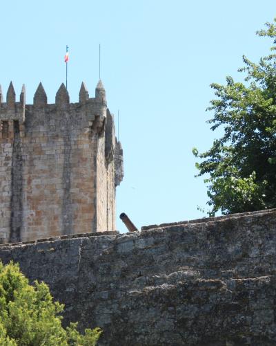 A stone medieval castle tower with crenellations and a Portuguese flag, situated behind a stone wall with a cannon, surrounded by green trees under a blue sky.