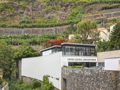 A modern white building with a glass upper level and a stone retaining wall, situated at the base of a steep, terraced hillside covered in vegetation, with the text "CENTRO CULTURAL JOHN DOS PASSOS" clearly visible on the facade.