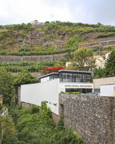A modern white building with a glass upper level and a stone retaining wall, situated at the base of a steep, terraced hillside covered in vegetation, with the text "CENTRO CULTURAL JOHN DOS PASSOS" clearly visible on the facade.