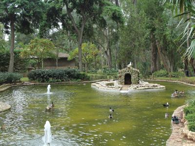 A large, irregularly shaped pond with fountains and swimming ducks, featuring a small stone grotto structure on a central island, surrounded by dense trees and manicured garden paths.