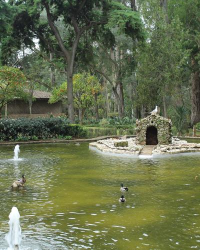 A large, irregularly shaped pond with fountains and swimming ducks, featuring a small stone grotto structure on a central island, surrounded by dense trees and manicured garden paths.