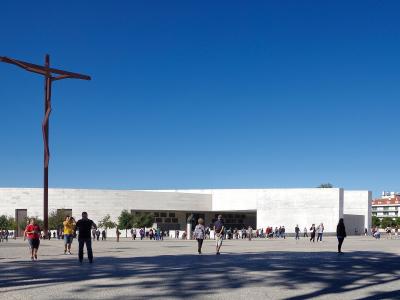 A wide shot of a large, modern white stone basilica with a tall, dark wooden crucifix sculpture standing prominently in the paved plaza in front of it under a clear blue sky.