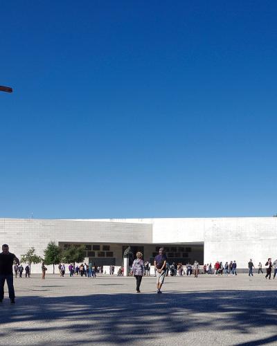 A wide shot of a large, modern white stone basilica with a tall, dark wooden crucifix sculpture standing prominently in the paved plaza in front of it under a clear blue sky.
