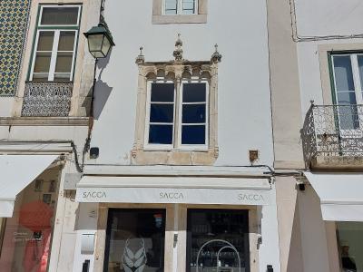 A close-up view of a white building facade featuring a distinctive Manueline-style window, with a shop named "SACCA" on the ground floor and outdoor seating.