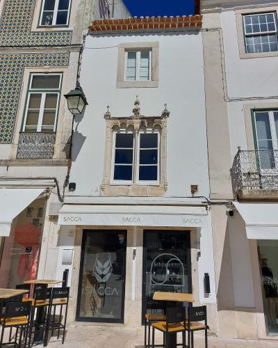 A close-up view of a white building facade featuring a distinctive Manueline-style window, with a shop named "SACCA" on the ground floor and outdoor seating.