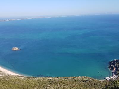 A high-angle panoramic view of a coastal bay featuring a sandy beach, turquoise water, a small rocky islet, and a distant sandbar, taken from a vegetated hillside.