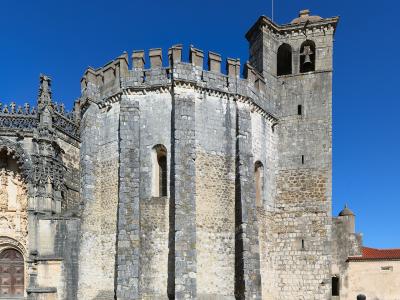 The image shows the exterior of a historic stone religious complex featuring a circular chapter house with crenellations, a bell tower, and an ornate Manueline-style portal on the left.
