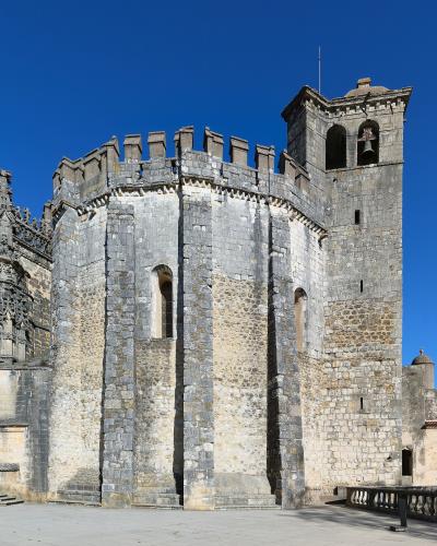 The image shows the exterior of a historic stone religious complex featuring a circular chapter house with crenellations, a bell tower, and an ornate Manueline-style portal on the left.