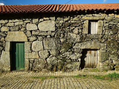 A close-up view of a rustic stone building facade with a terracotta tiled roof, featuring a green wooden door, a small shuttered window, and a lower wooden door, set against a cobblestone street.