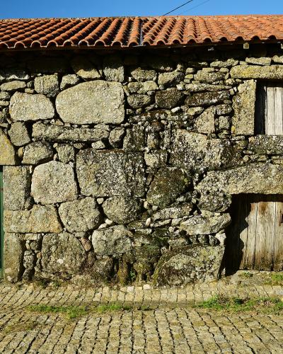 A close-up view of a rustic stone building facade with a terracotta tiled roof, featuring a green wooden door, a small shuttered window, and a lower wooden door, set against a cobblestone street.