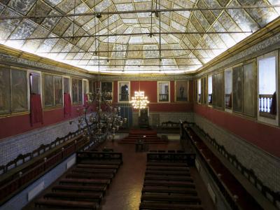 An interior view of a grand hall featuring a large glass and metal skylight ceiling, red walls adorned with portraits, blue azulejo tile wainscoting, and rows of wooden benches facing a raised dais.