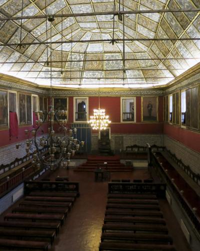 An interior view of a grand hall featuring a large glass and metal skylight ceiling, red walls adorned with portraits, blue azulejo tile wainscoting, and rows of wooden benches facing a raised dais.